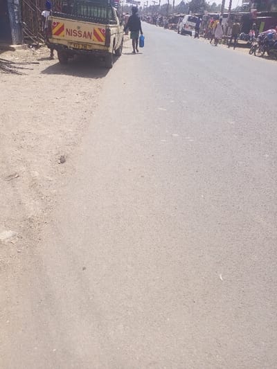 This image displays a street scene in Ongata Rongai, Kenya, observed from a low angle looking down a moderately wide, paved road that recedes into the distance. The immediate foreground on the left consists of a dry, unpaved dirt shoulder, while the right side is asphalt.
A cream-colored Nissan pickup truck with a green metal cage structure in its bed is parked on the dirt shoulder and partially on the paved road in the left midground, facing away from the camera. The truck's rear tailgate is visible, featuring "NISSAN" in prominent dark letters and a license plate reading "KVS 236". Red and yellow diagonal reflective tape is affixed to the tailgate's corners. A person's lower legs and feet are visible near the rear driver's side wheel of the truck, partially obscured.
Further down the paved road, a male individual with dark, curly hair, wearing a dark short-sleeved top and dark shorts, walks away from the camera on the paved surface, holding a blue plastic jerrycan or container in his left hand.
Along the right side of the road in the midground and background, numerous individuals are present, walking and standing. They are situated next to a line of structures that appear to be small shops or market stalls. Several vehicles, including a white van and at least two motorcycles, are visible parked along the roadside or in traffic further down the street. Utility poles and overhead wires are discernible against a clear sky in the distant background. The scene is brightly illuminated by natural sunlight, indicated by visible shadows.