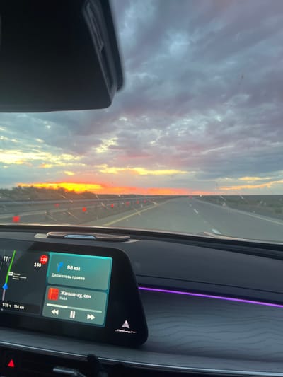 This is a first-person perspective, inside a car, looking out at a highway during sunset. The sky is filled with dramatic clouds, tinged with orange, yellow, and purple from the setting sun. A bright orange band of light is visible on the horizon. The highway stretches ahead, with red reflectors lining the right side. The car's dashboard is visible, featuring a digital display. The display shows navigation information, including a route with "98 KM" remaining and the instruction "Держитесь правее" (Keep right). It also displays music playback controls and song information for "Жаным-ау, сен" by RaiM. The car's interior has ambient lighting, with a purple light strip visible along the dashboard. The scene appears to be on a highway leading towards or away from the city of Жаркент, Kazakhstan, as indicated by the navigation display.