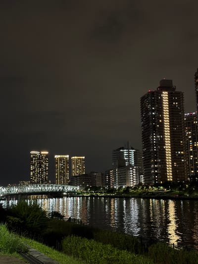 The image depicts a nighttime cityscape in Koto, Japan, centered on a wide river reflecting the illuminated buildings. On the left side of the river, a multi-span bridge with a white, latticed truss structure is visible, illuminated by lights along its length. The bridge connects to the distant shore, where several high-rise apartment buildings stand.

Across the river, on the right side of the frame, a dense cluster of modern, multi-story buildings dominates the skyline. The tallest building, in the mid-right, is a dark, slender skyscraper with a distinctive vertical band of bright yellow lights illuminating its central portion, suggesting it's lit from within. Numerous other windows in this and adjacent buildings are lit with a warm, yellowish glow. In the background, other residential towers and structures are visible, some with balconies.

The river itself is dark, with the lights from the bridge and buildings creating elongated, shimmering reflections that break up the surface of the water. The water appears calm, with gentle ripples disturbing the reflections.

In the foreground, along the left bank of the river, a grassy embankment slopes down towards the water. The grass is a dark green, with some patches appearing brighter where artificial lights might be present. A stone path or edge is visible at the very bottom left of the frame.

The sky above is dark and overcast, with subtle variations in shade indicating cloud cover. There are no discernible stars or moon. The overall mood of the image is one of urban nighttime serenity, with the man-made structures and their lights creating a vibrant contrast against the dark natural elements of the river and sky. No people are clearly visible in the scene.