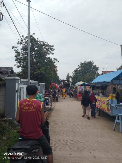 The image shows a street scene in Kecamatan Anjatan, Indonesia (latitude: -6.418, longitude: 107.969). The street is lined with houses, trees, and food stalls. A man wearing a red shirt with "BERKAH SWIKE ANAK WADON" written on it sits on a motorcycle facing away from the camera. Two women, one wearing a brown jacket and the other wearing a hijab, are walking down the street. A food vendor is standing behind a stall with a yellow and black sign. The street is made of concrete and has a dusty, uneven surface. There are power lines strung across the street. The sky is overcast.