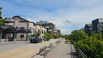 This image shows a street scene in the city of Linköping, Sweden, specifically in the Gottfridsberg district. The perspective is from a sidewalk looking down a street lined with modern apartment buildings and greenery.
In the foreground, there is a paved walkway with yellow and grey patterned tiles. Several wooden benches are placed along the side of the walkway. To the left of the walkway is a black electric car, a Renault Megane E-Tech, plugged into an electric vehicle charging station. Adjacent to the car is a covered structure with a sloped roof, possibly a bus shelter or a bicycle parking area, with glass doors and windows. Behind this structure, a pink, multi-story apartment building with a curved roof and black trim is visible.
Further down the street, a row of colorful apartment buildings with balconies and wooden facades follows. The buildings vary in height and architectural style, with some featuring metallic roofs. Trees and other greenery are interspersed between the buildings and along the sides of the street, creating a vibrant, residential atmosphere.
On the right side of the image, another paved walkway runs alongside a metal railing, bordering a dense area of green trees and bushes. The sky above is a bright blue with scattered white, fluffy clouds. The overall scene suggests a well-maintained, modern urban environment with a focus on sustainable living, given the presence of the electric car and charging station.