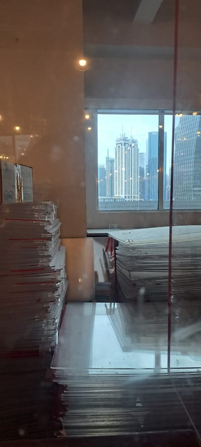 This is a rotated indoor photograph taken from a high vantage point, looking out a window. The scene is dimly lit with artificial lights reflecting off surfaces.

In the foreground, on the right side, stacks of transparent materials, possibly acrylic sheets or glass panels, are arranged vertically. Some of these materials have red vertical elements, possibly support or separators.

The middle ground shows a large window with a view of a city skyline. Several tall buildings are visible, including one with many horizontal lines and a distinctive crown-like structure at the top, which appears to be the Philippine National Bank (PNB) Financial Center. Another building, labeled "BDO," is also discernible. The sky is overcast.

The left side of the image features a wall with several circular light fixtures reflecting onto a smooth, likely polished surface. A red horizontal line is visible across the top portion of the image, potentially a structural element or part of the room's decor.

The overall impression is of an office or showroom environment, possibly during daytime given the ambient light from the window, but with artificial lighting also prominent. The arrangement of materials suggests a storage or display area. The presence of specific building names like "PNB" and "BDO" strongly suggests the location is in Metro Manila, Philippines, specifically consistent with the reported location of Mandaluyong, which is adjacent to the financial district where these buildings are located. No people are visible. The weather appears to be cloudy.