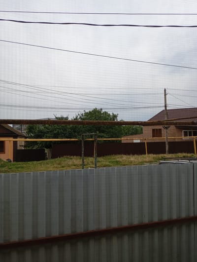The image depicts an outdoor scene with a focus on utility infrastructure and residential structures in Shelkovskaya, Russia. A grey corrugated metal fence occupies the foreground, with a rusty metal beam running horizontally across its lower section. Behind this fence, a grassy area is visible, transitioning into a brown fence.
Beyond the brown fence, a large, rusted metal pipe runs horizontally across the mid-ground. To the left of this pipe, a portion of a small, light-colored building with a brown roof is visible. In the center and right mid-ground, a brick building with a brown tiled roof and wooden-framed windows is present, along with a tall, light-colored utility pole supporting multiple overhead wires. Dense green trees are situated behind the fences and between the buildings.
The sky above is overcast with grey clouds, and a network of power lines crisscrosses the upper portion of the image. The overall impression is of a somewhat utilitarian and rural or semi-urban environment.