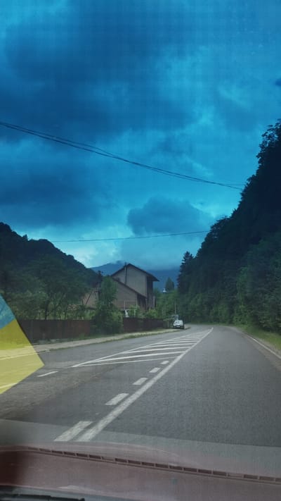 The image depicts a view from inside a vehicle, likely a car, on a road near Rakhiv, Ukraine. The right side of the image shows an asphalt road with white lane markings, bordered by green vegetation. A vehicle is parked alongside the road. On the left side, the view is dominated by a blue sky with dark cloud cover, possibly reflecting in a windshield. A building is visible amidst the greenery, further away from the road. Overhead power lines are also present. The image is slightly rotated, with the road oriented vertically.