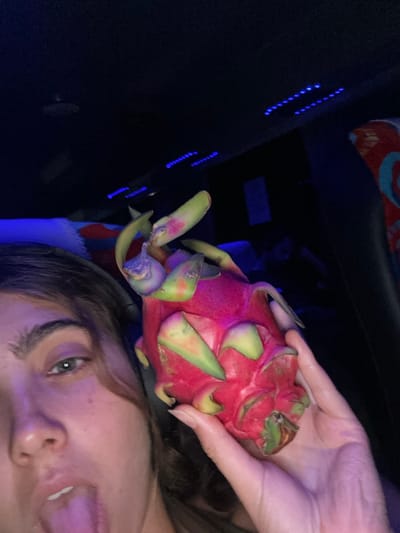 The scene is set inside a vehicle in Lang Suan, Thailand. A female subject is partially visible on the left side of the frame, showing her face from the chin to just above the right eye. She has dark hair and a visible eyebrow; her tongue is extended from her mouth, pointing downwards and slightly to the left. Her right hand is holding a large, ripe dragon fruit with vibrant pink skin and green-tipped bracts, positioned in the central-right portion of the image. The immediate background is dark, revealing multiple blue LED strip lights on the vehicle's ceiling and a section of a multi-colored patterned fabric, likely a seat or upholstery, on the far right.