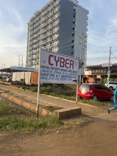 A sign advertises "CYBER" services including printing, photocopying, business cards, binding, lamination, scanning, KRA PIN & registration, filing returns, Wi-Fi subscription (20 Mbps), surfing, office space, and passport photos. Another sign indicates "KHARM WASH WORLD SERVICES" is nearby. In the background, a tall, modern building rises behind power lines, and cars are parked on the street. The image appears to be taken on a sunny day.
