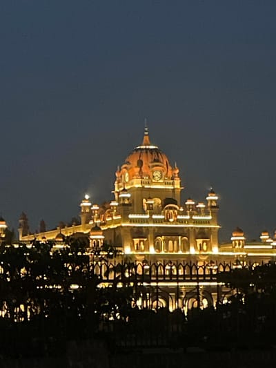 The image shows a large, illuminated building at dusk or night. The building features multiple domes, ornate architecture, and is lit with warm-toned lights, highlighting its details. A fence and dark foliage are in the foreground, obscuring the lower part of the building and suggesting a viewpoint from across an open area. The sky is a deep, dark blue, indicating twilight or night. This building is the Rambagh Palace in Amritsar, India, a former royal residence and now a heritage hotel. There are no people visible in the image.