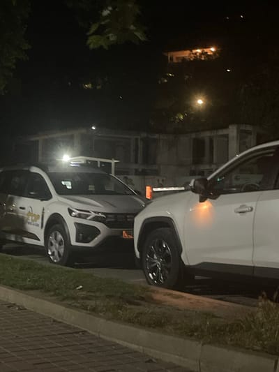 The image displays two white vehicles parked side by side at night in what appears to be an urban or suburban setting. The vehicle in the foreground has its driver's side door open, revealing a glimpse of the interior. The vehicle behind it is also white and appears to be a similar model. Both vehicles have their headlights off. The ground is paved with bricks, and there is a strip of grass and a curb visible. In the background, there is a building with multiple windows. The lighting suggests it is nighttime, and the scene is illuminated by streetlights or lights from the building. There is some foliage visible in the foreground, suggesting the presence of trees or bushes. The overall mood is quiet and still, typical of a nighttime scene. There is some text visible on the side of the vehicle in the foreground, but it is too blurry to read clearly. The image seems to be taken from a slightly elevated angle, looking down at the vehicles. No people are visible in the frame. The crops highlight specific details like the car doors, wheels, and the surrounding environment.