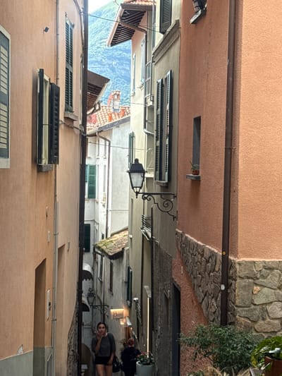 The image depicts a narrow, slanted street in San Giovanni, Italy. Buildings with textured peach and gray facades line both sides of the street, featuring dark-shuttered windows. A quaint lamp post is mounted on a building on the right. In the background, lush green mountains rise, partially obscured by the buildings.
A young woman with dark hair, wearing a gray top and black shorts, is walking down the street, holding something to her mouth. Another person, with red hair and wearing a dark jacket, is visible further down the street. The ground is paved with cobblestones. The scene is illuminated by natural daylight, creating a somewhat shadowed atmosphere due to the narrowness of the street.