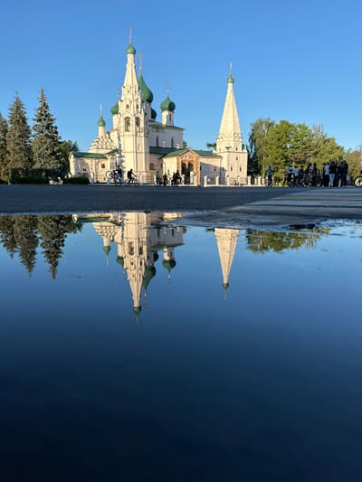 The image depicts a reflection in water, creating an upside-down view of a scene. The primary subject is a white church with green domes, characteristic of Russian Orthodox architecture. It is a sunny day, and the bright blue sky is visible. Trees with lush green foliage surround the church.
In the foreground, which is the reflection, a paved area with some wet patches suggests recent rain or a damp surface. Several bicycles are parked in a row. A group of people, mostly dressed in casual attire, are visible near the bicycles and the church. Their presence suggests a public space, possibly a tourist attraction or a gathering point.
The reflection creates a striking visual effect, with the church and trees appearing to float in the dark, reflective surface which dominates the left side of the image. The light conditions suggest it is daytime, likely morning or afternoon, given the clear sky and bright lighting. The overall mood is serene, with the reflection adding a touch of artistic or dreamlike quality to the scene. There is no visible text in the image. The setting is consistent with a location in Yazvitsevo, Russia, given the architectural style and natural environment.