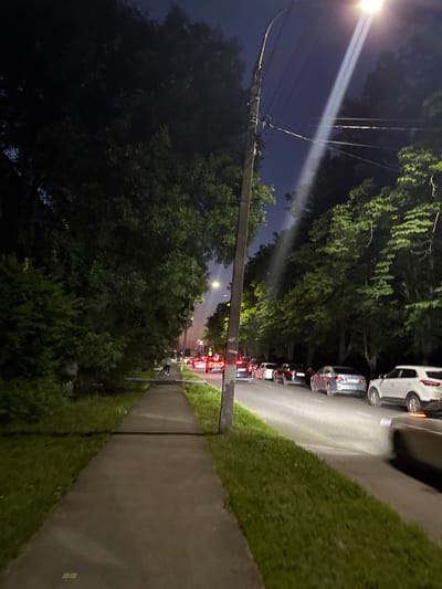 The image depicts a street scene at night, likely in a residential area with trees lining the road. A lamppost shines a bright beam of light upwards, cutting through the dark sky. On the right side of the road, there is a sidewalk and a grassy area. Several cars are parked or driving on the road, with their headlights and taillights illuminated, suggesting traffic. The overall mood is calm and quiet, typical of a late-night setting. There is no visible text in the image.