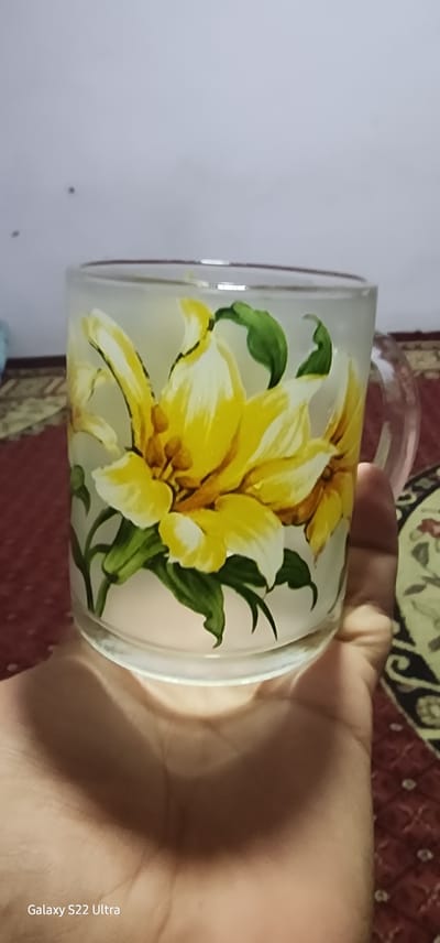 A hand holds a frosted glass mug decorated with yellow lilies and green leaves. The mug is clear glass with a frosted effect, and the floral design is painted on the exterior. The background consists of a red patterned carpet and a plain wall. A watermark at the bottom left reads "Galaxy S22 Ultra". The image appears to be taken indoors.