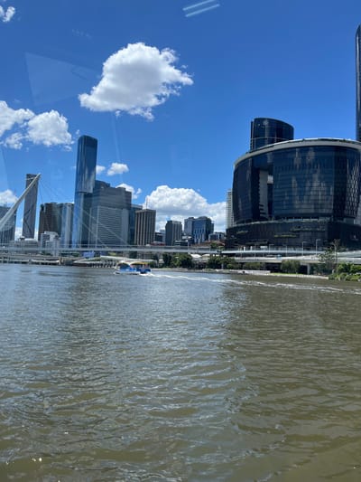 The image presents a vibrant urban cityscape under a clear blue sky, viewed across a wide river. The perspective is from an elevated position, looking towards the city skyline.

The foreground and midground are dominated by a broad, calm river, likely the Brisbane River, characterized by its brownish-green water with numerous ripples and sparkling reflections from the bright sunlight. A few small watercraft, possibly recreational vessels, are visible on the water. Along the riverbank, a long, paved promenade or walkway can be seen, with a few small, indistinct human figures strolling or standing.

The city skyline rises majestically on the far side of the river. A prominent feature is a large, curved building with dark, reflective glass panels and white trim, possibly a convention center or a significant corporate building. A subtle white logo, resembling a stylized floral or star shape, is visible on one of its facades. Beyond this, numerous other modern high-rise buildings and skyscrapers form a dense urban environment, showcasing varied architectural designs and reflective surfaces that mirror the sky and surrounding structures. A tall, slender skyscraper is particularly noticeable further along the bank. Elements of bridges, including visible suspension cables and structures, connect different parts of the cityscape.

The sky above is a brilliant azure, sparsely dotted with fluffy white cumulus clouds. The bright conditions indicate a sunny day, likely midday or early afternoon, with strong natural light illuminating the entire scene. The overall atmosphere is one of serene urban activity, with the modern architecture contrasting with the natural elements of the river and sky. No specific text is clearly legible on any structures beyond the logo mentioned.
