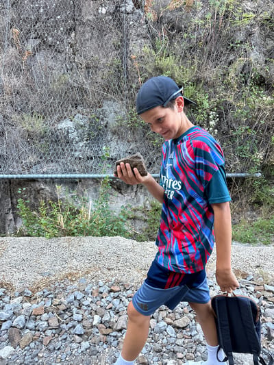 A young boy wearing a baseball cap, a colorful patterned t-shirt, and blue shorts walks along a rocky path. He is holding a rock in his right hand and looking down at it. A black backpack is slung over his left shoulder. Behind him, a rock face is covered with a wire mesh fence, and various plants and trees are growing. The ground is covered with gravel and stones. The scene appears to be outdoors in a natural setting, possibly a hiking trail or mountainous area.