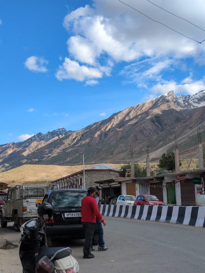 In the city of Padum, India, a mountain landscape is visible with snow-capped peaks under a blue sky dotted with white clouds. In the foreground, a road runs alongside a row of buildings. Two men are standing on the road near several vehicles, including a silver pickup truck, a black car, and a scooter. A white car and a red car are parked in front of the buildings. The buildings appear to be shops, with signs indicating "Lamo Fancy" and "Oppo." The scene suggests a street in a town situated in a mountainous region.