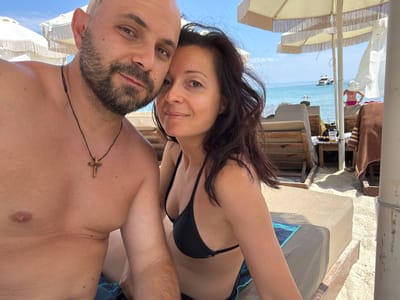 The original image is a selfie taken by a couple on a beach. The man on the left has a shaved head and a beard, wearing a black necklace with a wooden cross. The woman on the right has dark, long hair and is wearing a black bikini top. They are both smiling and looking at the camera. Behind them, beach chairs and umbrellas are visible, as well as the turquoise sea and a white yacht in the distance. The scene suggests a relaxed holiday atmosphere. The location appears to be a beach in the Pallini Municipal Unit of Greece, likely a popular tourist area given the presence of beach amenities and a yacht.