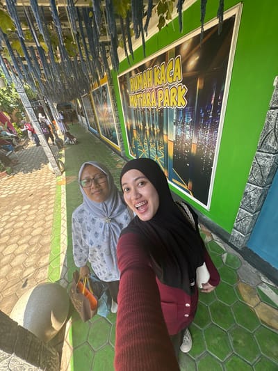 Two women are taking a selfie in front of a building with colorful murals and a sign that reads "RUMAH KACA MUTIARA PARK" in Tegalwangi, Indonesia. The woman on the left is wearing glasses and a light blue hijab, while the woman on the right is wearing a black hijab and a maroon sweater. Both women are smiling. The ground is paved with hexagonal tiles, and there are plants and hanging decorations overhead. The overall scene appears to be a tourist attraction or entertainment venue.