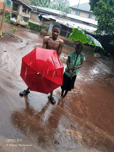 This image captures two young individuals caught in a downpour in Akokwa, Nigeria. A teenage boy, shirtless and wearing a necklace, stands on the left, holding a large, closed red umbrella. To his right, a younger child, dressed in a patterned blue and green shirt and a black skirt, uses a large green leaf as a makeshift umbrella.
The ground is a muddy brown, slick with rain and reflecting the surrounding environment. Water is clearly pooling and flowing, indicating heavy rainfall. In the background, there are glimpses of rustic structures, including what appears to be a chicken coop with wire mesh, and a corrugated metal roof. Further back, a two-story building with visible windows can be seen. Lush green foliage surrounds the scene, typical of the tropical climate.
The lighting suggests it is daytime, though the overcast sky due to the rain likely diffuses the light. The overall mood is one of resilience and perhaps a bit of playfulness amidst the challenging weather conditions. The presence of water and mud, the makeshift umbrella, and the children's attire all point to a rural or semi-rural setting in Nigeria. Visible text at the bottom of the image reads "OPPO R11" and "Nigeria Obodoukwu", confirming the location.