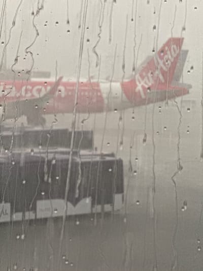 An AirAsia airplane is visible through a window covered in water streaks and droplets. The airplane is predominantly red with white lettering that reads "Air Asia" on its tail. In the foreground, partially obscured by the water, is a section of a bus or service vehicle, which appears to be dark purple with white markings. The scene suggests an airport environment, possibly during rainy weather, given the water on the window. The overall image is blurry and desaturated due to the rain and the viewing angle. The location is described as Nong Prue Subdistrict, Thailand.