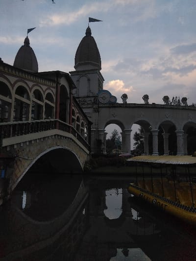 The image displays an architectural complex featuring prominent domes and arches, situated alongside a body of water in Ciudad de San José, Costa Rica. The sky above is partially cloudy, indicating twilight conditions.
In the left foreground, a building with a dark, ridged dome topped by a dark pennant flag is visible. An arched bridge, constructed from light-colored stone with a dark-topped railing, extends from this building, spanning the dark, still water. The bridge's arch and the building's lower section are reflected on the water's surface.
Further to the right and slightly behind, a taller structure with an identical dark, ridged dome and flag rises. To its right, a long, horizontal facade features a series of rounded arches supported by columns, with ornamental urns or finials spaced along its upper edge.
In the lower right corner, a yellow boat with a dark canopy is moored on the water. The boat's visible side features the text "BAKI BOAT" in dark letters, bordered by a dark checkered pattern. Reflections of the boat and the distant arched structures are visible in the water. No individuals are present in the scene.