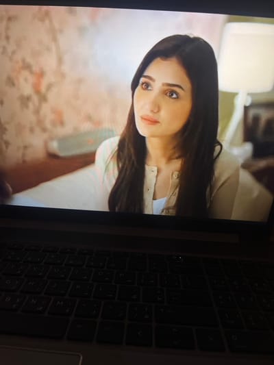 The image displays a close-up of a laptop screen showcasing a young adult female from the chest up, situated in an indoor environment. The woman has long, dark brown hair and is wearing a light beige or cream-colored button-up cardigan over a white top. Her face, in profile facing slightly left of center, shows a contemplative expression with her gaze directed upwards and to the right. The background on the screen features a wall with light-colored floral wallpaper on the left, and a portion of a white table lamp is visible on the right. Below the screen, the laptop's keyboard is visible, with black keys displaying both Arabic and English characters, dimly illuminated. This scene is captured in Nuseirat, Palestinian Territory.
