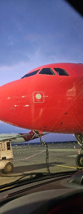 The scene depicts the nose and cockpit section of a bright red narrow-body passenger aircraft, likely an Airbus A320 series, situated on an airport apron at Katowice Airport (KTW) in Pyrzowice, Poland. The sky overhead is a deep blue, indicative of twilight. Artificial lighting on the apron casts reflections on the aircraft's red fuselage. A circular sensor is visible on the forward fuselage below the cockpit windows. To the left and rear of the red aircraft, the tail and a segment of a second, white aircraft with red livery are discernible. A white ground support vehicle, equipped with yellow wheels, is positioned on the apron to the lower left. The immediate foreground reveals the dashboard and windshield wipers of the vehicle from which the image was captured. The ground surface is asphalt or concrete, featuring yellow and white demarcation lines, with orange cones visible in the distance. No individuals are explicitly present.