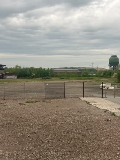 The image depicts an industrial landscape near Schifflange, Luxembourg, characterized by a large, spherical gas holder in the distance. The foreground consists of a gravel area bordering a concrete structure, with a black metal fence running along the perimeter. Beyond the fence, there is vegetation including trees and shrubs, leading to further industrial structures. Power lines can be seen in the midground. The sky is overcast with heavy clouds. There are no people visible in the image. The scene appears deserted or disused.