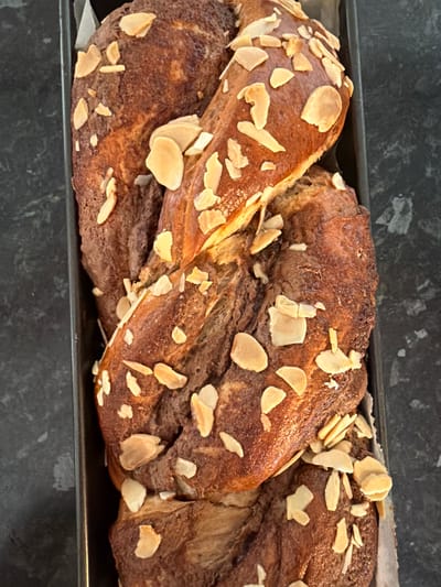 A freshly baked, almond-topped braided bread loaf sits in a dark metal baking pan. The loaf has a dark brown, slightly mottled crust and is generously sprinkled with almond slices. The pan rests on a dark, possibly granite or composite, countertop. The scene appears to be indoors, likely in a kitchen. There is no indication of the location being in Bretton, United Kingdom, or of any people.