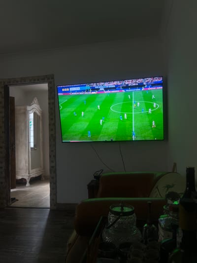 This is a photograph taken indoors, likely in a living room or entertainment area in Brighton, United Kingdom. The primary subject is a large flat-screen television mounted on a wall, displaying a soccer (football) match. The game appears to be a broadcast from the FIFA World Cup 2026. Several players in white and blue uniforms are visible on the green field, with spectators in the background. Text on the screen indicates the broadcast is from BBC, with the score at 0-0 between ENG (England) and DRC. The time displayed is 03:16. The television is positioned above a mirrored archway, which reveals a reflection of a room with ornate furniture, possibly an antique cabinet or wardrobe. To the right of the television, a shelf holds several bottles, including what appears to be wine, and glassware, suggesting a bar area or a place where drinks are prepared or displayed. The lighting in the room is dim, with a spotlight or ray of light illuminating the floor on the right side of the image. The overall scene conveys a relaxed, domestic atmosphere, perhaps someone enjoying a sporting event at home.