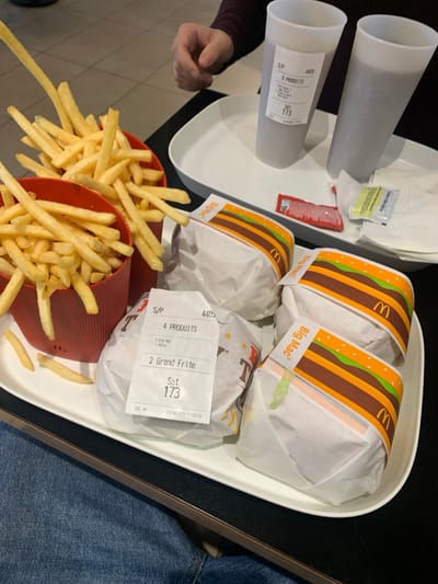 The image depicts a meal served on a dark table within a McDonald's restaurant located in Fontenay-sous-Bois, France, dated October 23, 2025, at 11:23:16. A large white rectangular tray holds two red plastic containers brimming with French fries, one fry extended vertically as if being picked up. Four wrapped burgers are positioned next to the fries; three are identified as "Big Mac" by their branded wrappers. The fourth burger, wrapped in plain white paper, has an attached receipt detailing the order: "4 PRODUITS", "3 Big Mac", "1 Tasty", and "2 Grand Frites". A smaller white tray holds two translucent white plastic cups containing a dark beverage, each with a receipt affixed. Two red ketchup packets and one yellow-green condiment packet rest on this tray. A human right hand and forearm, clad in a dark long-sleeved garment, are visible on the right side of the frame, near the drinks. Partially visible at the bottom left are the legs of another individual, wearing blue jeans. The background shows light-colored tiled flooring.