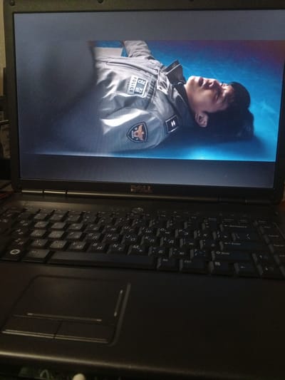The image shows a close-up of a laptop screen displaying a scene from a video. On the screen, a person is lying on their back, wearing a grey uniform with a patch that reads "POLICE" and Korean characters. The person's eyes are open, and they appear to be in distress or unconscious. The background on the screen is a bright blue surface. Below the screen, the keyboard and touchpad of the black laptop are visible. The keyboard appears to have a Cyrillic layout. The image does not provide any information about the city of Yessentukskaya, Russia, or any specific location.