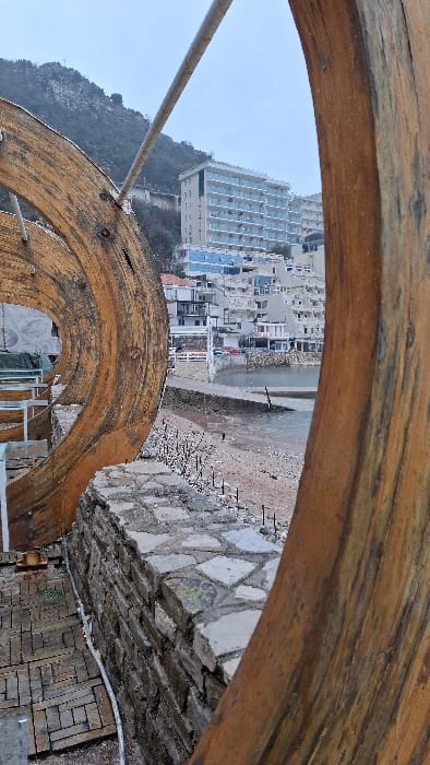 The image presents a view of Podbadac, Montenegro, framed by two large, weathered, curved wooden structures. In the immediate foreground on the left, a section of a wooden planked platform is visible, with a white cable running along its surface. A stone wall, constructed from irregular grey and brown blocks topped with flatter flagstones, extends horizontally across the lower midground. Beyond this wall, a narrow sandy and pebbly beach leads to a body of water. A concrete pier or ramp extends into the water from the left side of the frame. Along the coastline, a cluster of buildings with white and blue facades is visible, with a red vehicle parked near the pier. In the background, ascending a hillside, are more buildings, including a prominent multi-story structure with numerous balconies. A steep, rocky mountain with sparse vegetation rises in the far distance under an overcast sky. A metal rod or pipe stretches horizontally across the upper portion of the frame, intersecting with the framing wooden structures. No individuals are visible in the scene.