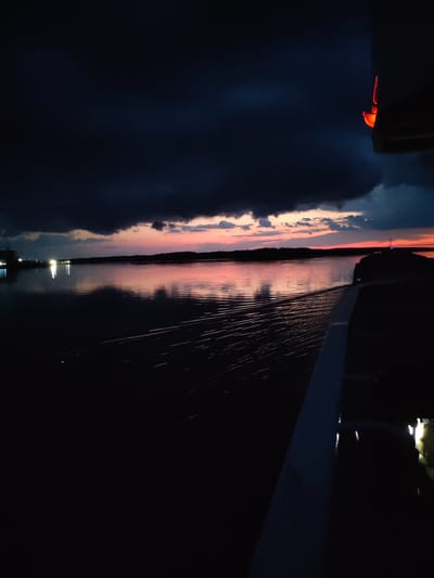 This media file captures a scenic twilight view of a body of water, likely a river or lake, near Bol'shie Mkhi, Russia. The sky is a dramatic canvas of dark, heavy clouds transitioning into a soft pink and orange sunset glow near the horizon. The water reflects the colors of the sky, creating a shimmering, tranquil surface.
The scene is viewed from the perspective of a moving vessel, possibly a boat or ferry, as evidenced by the wake trailing behind and the railing of the vessel visible in the foreground. The dark silhouette of a distant shoreline is visible, dotted with lights that suggest a settlement or structures. The overall atmosphere evokes a sense of calm and peaceful transition from day to night.
The time of day is clearly twilight or dusk, with the fading light creating a moody and atmospheric setting. The weather appears to be overcast, with thick clouds dominating the upper sky. The location cues, such as the distant lights and the broad expanse of water, are consistent with a riverside or lakeside environment in a rural or semi-rural area.
There are no discernible people or specific objects of focus, other than the natural landscape and the moving vessel. The focus is on the interplay of light, water, and sky during a beautiful sunset. The emotions conveyed are primarily serenity, tranquility, and a sense of wonder at the natural beauty of the scene. There is no visible text in the image.