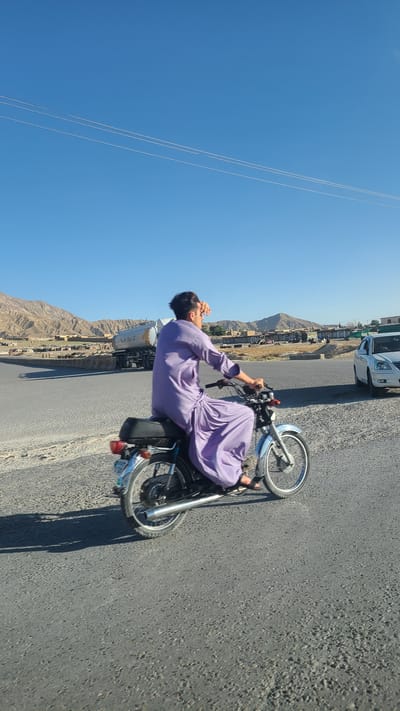 The image depicts a man riding a motorcycle on a paved road. The man is dressed in a light purple robe. He is touching his face with his hand. The motorcycle is silver and black.

The background shows a clear blue sky with power lines, a mountain range, and buildings indicating a populated area. There are other vehicles on the road, including a white car and a tanker truck. The location is Kuchlak, Pakistan.