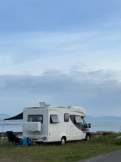 This media file shows a white Autotrail motorhome parked on a grassy verge next to a road, overlooking a body of water. The motorhome has the model name "HIMALAYA" visible on the back. There is a small awning extended from the side of the motorhome, and two camping chairs are set up underneath it. One chair is occupied by a person wearing a dark top. A few bags and towels are also visible near the chairs.

The scene appears to be a coastal or lakeside setting, with hills or mountains visible in the distance across the water. The sky is partly cloudy, with patches of blue and white. The overall impression is one of a peaceful outdoor scene, likely during daytime. The image is oriented vertically, suggesting it might have been taken on a phone and rotated. The license plate of the motorhome reads "CGZ 8358". The location context provided is Terhillion, Ireland.
