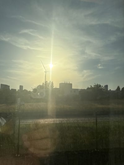 This media file captures a scene during what appears to be late afternoon or early evening, given the bright, low-hanging sun. The sky is filled with wispy, streaky clouds, suggesting a partly cloudy or hazy condition. The sun is a dominant feature, creating a lens flare that extends across the image, indicating it is either directly in view or very close to the camera.
The primary object in the landscape is a large wind turbine, its blades visible against the bright sky. In the middle ground, there are several silhouetted buildings, appearing to be low-rise structures and possibly some taller buildings in the distance, interspersed with trees. The overall setting seems to be a semi-rural or suburban area. A fence line runs parallel to the right side of the frame, separating the foreground from a grassy or vegetated area. The ground appears to be covered in dry grass and weeds. There are some indistinct signs visible near the fence line. The overall mood is somewhat serene, with the strong sunlight casting a golden hue over the landscape. The image appears to have been taken from a moving vehicle, possibly a car, given the slight blur and the reflective surface on the right side of the frame, which may be a window. There is no discernible text in the image.