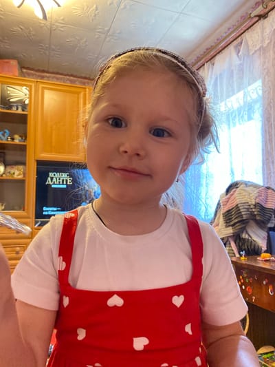 A young girl with blonde hair and blue eyes, wearing a white t-shirt and a red dress with white hearts, is pictured from the chest up. She has a headband with rhinestones in her hair. The background includes a wooden cabinet, a television displaying text in Russian that reads "КОДЕКС ДАНТЕ" (Dante's Code), and a window with lace curtains. A striped blanket is draped over furniture to the right. The scene appears to be indoors, possibly a living room. The location is identified as Mariupol, Ukraine.