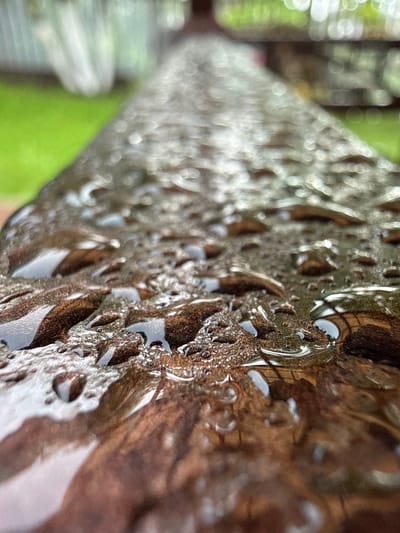 The image is a close-up, low-angle shot of a wet wooden surface. The wood appears to be dark brown and has a rough texture, with numerous raindrops clinging to its surface. The raindrops are of varying sizes and are reflecting the surrounding environment, creating bright highlights and distorted images within them. The background is blurred, but hints of green grass and light-colored, possibly white, blurry objects suggest an outdoor setting. The perspective draws the viewer's eye along the length of the wooden surface, which stretches into the distance, creating a sense of depth. There are no people or animals visible in the image. The scene appears to be one of a natural environment after rain.