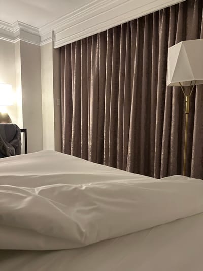 A room's interior features a unique ceiling design with horizontal, textured panels in a muted brown tone, illuminated by a geometric white lamp. A white curtain or fabric hangs prominently, partially obscuring the view, with a glimpse of a person or object beneath it. The wall is painted in a neutral color, and ornate white molding is visible along the top edge of the wall.