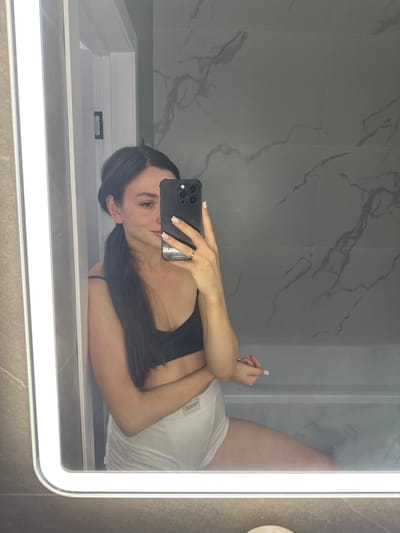 A woman is taking a mirror selfie in a bathroom. She has dark hair pulled back into a ponytail and is wearing a black bra and white shorts. She is holding a black smartphone in her right hand, angled to capture her reflection. The bathroom features marble tile walls and a bright, illuminated mirror. The woman appears to be leaning against a doorframe, with her left arm crossed over her chest and her right arm extended to hold the phone. The reflection shows her face, upper body, and legs. There is no visible indication of the city of Estachi, Russia.