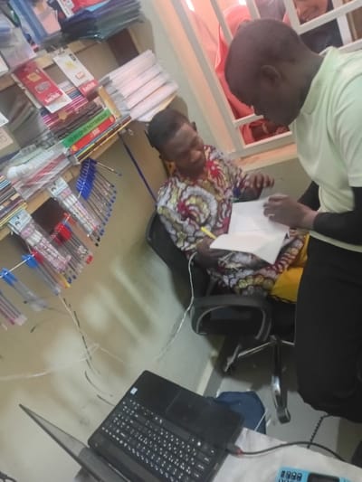 This image depicts an indoor commercial or office setting in Kwazailewa, Nigeria, characterized by stationery supplies. Two adult males are engaged in a transaction or consultation, while a third individual is partially visible in the background.
The male on the left is seated in a black office chair, wearing a multicolored patterned long-sleeved shirt. He holds a yellow pen or marker in his right hand, looking intently at a stack of white papers held by the second male, with a slight smile. The male on the right is standing or leaning, dressed in a light green short-sleeved top worn over a dark long-sleeved undershirt. He holds a stack of white papers, appearing to present or review them with the seated individual, his right hand positioned over the documents.
In the foreground, a black laptop is open on a reflective surface, with a red USB drive or connector inserted into its right side. A light blue keypad mobile phone is positioned to the right of the laptop, connected by a white cable.
To the left, a multi-tiered wooden shelving unit displays various stationery items. Visible products include packages of SanDisk memory cards, stacks of notebooks, and numerous pens of assorted colors (blue, red, black) hanging from hooks. The walls are light-colored. Several white electrical cables are visible, traversing the wall and floor. In the upper right background, a white-barred window or partition reveals the upper portion of a third individual, partially obscured, wearing what appears to be a light pink head covering.