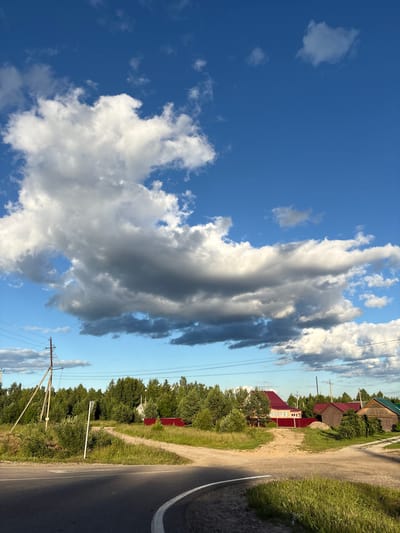 The image shows a rural scene in Kady, Russia. The sky is a clear blue with scattered white and gray clouds. A road, partially paved and partially dirt, runs along the right side of the frame, bordered by green grass and trees. Several houses with red and wooden roofs are visible among the trees. Power lines and poles are also present in the scene. The lighting suggests it is late afternoon or early evening, with the sun casting a warm glow. The overall atmosphere is peaceful and serene. There is no visible text in the image.