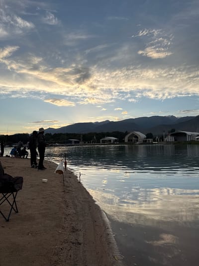The image depicts a serene lakeside scene at sunset in Cholpon-Ata, Kyrgyzstan. The foreground shows a sandy beach with several people engaged in fishing. Two figures stand in silhouette against the dramatic sky, holding fishing rods. A fishing net is visible on the sand, along with a foldable chair. Further down the beach, other individuals can be seen fishing or resting.

The lake occupies the right side of the frame, its surface reflecting the warm hues of the setting sun and the wispy clouds in the sky. The water appears calm, with gentle ripples distorting the reflections. In the background, silhouetted mountains rise behind a line of trees and some modern architectural structures, possibly part of a resort or residential area. The sky is filled with an array of cirrus clouds, illuminated by the golden light of dusk, creating a visually striking contrast with the deep blue of the sky.

The time of day is clearly sunset, indicated by the warm lighting and long shadows. The weather appears to be clear and pleasant, conducive to outdoor activities. The overall atmosphere is peaceful and relaxed, suggesting a moment of quiet enjoyment by the water. No visible text is present in the image.