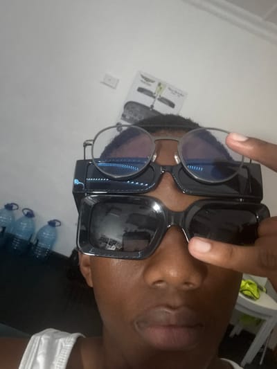 The image depicts a close-up, upward-angled view of a person's upper face and head, located indoors in Port Alfred, South Africa. The individual, of indeterminate gender and with dark skin, faces the camera while wearing three distinct pairs of glasses stacked vertically on their face. The topmost pair consists of round, metal-framed glasses with clear lenses exhibiting a blue light reflection. Directly beneath, a pair of black, rectangular glasses, possibly smart glasses, is worn, also showing blue internal reflections. The lowest pair comprises large, rectangular, black-framed sunglasses with dark lenses. The person's right index finger is positioned against the right side of the uppermost glasses' frame. A white sleeveless garment is visible at the bottom left of the frame. The background consists primarily of a plain white wall. To the left, a dark surface holds at least four translucent plastic water bottles with blue caps. On the wall, above the bottles, a white rectangular electrical fixture is visible. Further right, a paper document or poster depicting an aircraft, possibly a biplane, with accompanying text, is affixed to the wall. A fragment of a white table and a yellow-green fabric item are discernible on the far right.