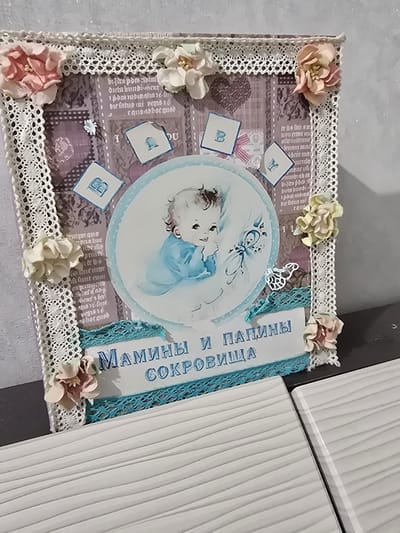 This media file shows a close-up, top-down view of a decorative box, likely a gift or keepsake, with a handmade aesthetic. The primary subject is the box itself, which is adorned with a variety of embellishments. There is a circular illustration in the center, featuring a baby in a blue outfit, possibly representing a newborn or young child. Surrounding the illustration are decorative elements like lace trim, artificial flowers in soft pinks and creams, and small, square tags with blue letters, one of which appears to be a "Y" and another a "B".
The box's surface is covered in patterned paper that has a vintage or scrapbook feel, with faint text and designs that are mostly illegible in the image. A prominent blue ribbon or trim runs along the right side of the box, bearing Russian text that translates to "MOMMY AND DADDY'S TREASURES." The overall presentation suggests a sentimental item, likely related to a child's birth or early life.
The setting appears to be indoors, with a neutral, textured surface beneath the box. To the right of the box, two white, ridged surfaces are visible, possibly parts of furniture or a wall, indicating a domestic or crafting environment. There are no clear indicators of time of day or weather, and the mood conveyed is one of affection and remembrance due to the baby imagery and the text. The location context provided is Satbayev, Kazakhstan, which doesn't offer specific visual cues within the image itself.