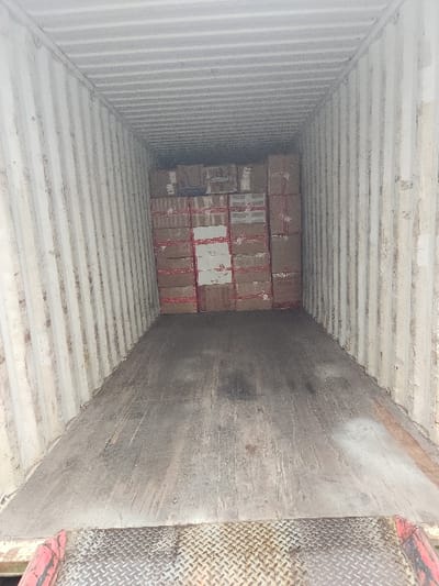 The image displays the interior of a weathered cargo container, located in Lakewood, United States. The container's corrugated metal walls and ceiling are a dirty off-white, exhibiting wear, streaks, and discoloration. The floor is a dark, grimy wooden surface, showing scuff marks, dirt, and minor debris. At the far end of the container, numerous uniformly sized brown cardboard boxes are stacked tightly, filling the space from floor to near-ceiling and wall-to-wall. Many boxes are secured with red packaging tape applied in horizontal and vertical configurations, and some feature white labels or markings. The immediate foreground includes a section of the worn wooden floor and a metallic diamond plate threshold. No individuals or active operations are visible within the container.