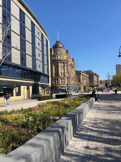 The image shows a street scene in Bradford, UK, located at 53.793 latitude and -1.753 longitude. The scene is captured from the perspective of a person standing on a paved sidewalk with a stone retaining wall to the left and a cobblestone path extending to the right. The sidewalk is lined with green vegetation and trees. In the distance, a row of buildings is visible. The tallest building is a large, ornate structure with a dome and a brown copper roof. The building is made of light brown stone and has numerous windows. The other buildings in the row are similar in style but smaller in height. There are people walking along the sidewalk, and one person is sitting on a bench on the right side of the image. The sky is blue and clear, and the sun is shining. The scene is typical of a bustling city street with a mix of modern and historic architecture. A light pole is visible in the distance at the end of the path. There are two people on the path, one is walking and the other is sitting. Several people are walking in the distance. The scene is captured in a clear, sunny day with well-lit conditions.