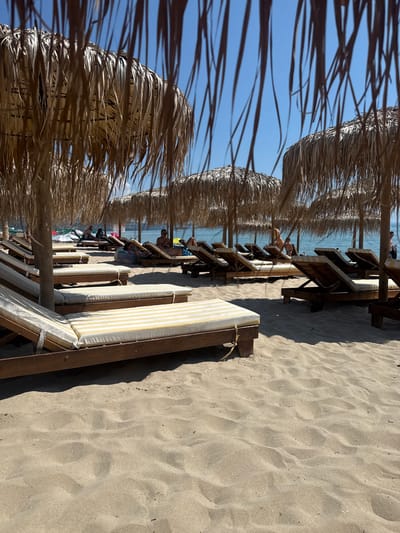 The image shows a beach in Kranevo, Bulgaria. The foreground is dominated by sand and several empty, sun-bleached lounge chairs with yellow and white striped cushions. Behind the chairs, more lounge chairs and people are visible, some reclining and some standing. Numerous straw umbrellas provide shade over the lounge chairs. The ocean is visible in the background, with clear blue water. The sky is also a clear, bright blue, suggesting a sunny day. The overall scene depicts a typical resort beach setting.
