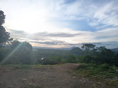 The image displays an elevated landscape view located in Geundring, Indonesia. The upper two-thirds of the frame are dominated by the sky, transitioning from an overexposed, bright white area with a pronounced sun glare on the upper left to a light blue expanse with scattered white and grey stratocumulus clouds across the right.
The immediate foreground consists of unpaved ground, primarily composed of light brown dirt and gravel. This ground features intermittent patches of green grass and other low-lying vegetation, along with scattered pieces of white debris.
The midground is characterized by a dense, irregular canopy of green trees and bushes. The foliage on the left side of the frame appears partially silhouetted due to the bright sun glare. Embedded within the distant midground greenery, faint outlines of structures with visible roofs are discernible.
The background presents a panoramic vista of multiple rolling, tree-covered hills and mountains, receding into a hazy horizon. A distinct, steeply sloped, tree-covered mountain peak is prominently located in the mid-right section of the background. No human figures or specific activities are observed within the scene.
