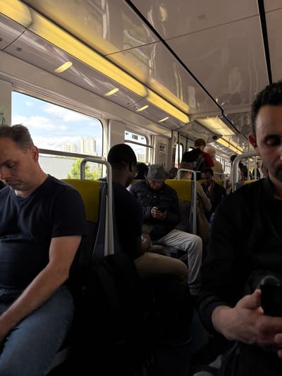 The interior of a train, likely a commuter train, in the city of Bourg-la-Reine, France, at the location with coordinates 48.785, 2.313. The scene is dimly lit, with overhead fluorescent lights providing the primary illumination. The train is relatively empty. A man in a black t-shirt sits in the foreground, looking down. Behind him, to the right, another man in a dark jacket sits with his hands in his lap. The second man has his back to the camera. The train is moving, as evidenced by blurred images outside the window. A man in a blue jacket and white pants is seated across from the man in the black t-shirt. He is looking down at his phone. A man in a red shirt is standing near the back of the train, likely talking to another person out of frame. There is a window view of buildings outside the train. The air conditioning vents are visible above the seats. Several passengers are wearing headphones. The train is clean and appears to be relatively new.